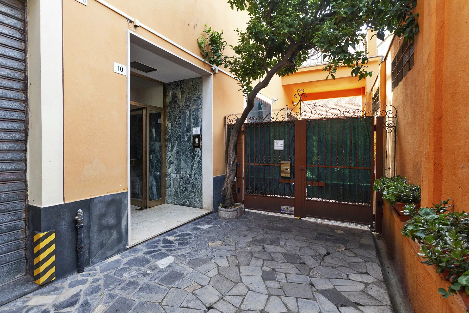 Un patio de entrada con una puerta metálica, pavimento de piedra y un árbol.