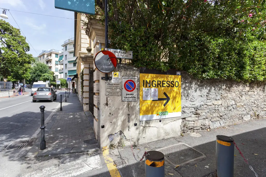Una calle urbana soleada pasa junto a un muro de piedra con un letrero amarillo de entrada.
