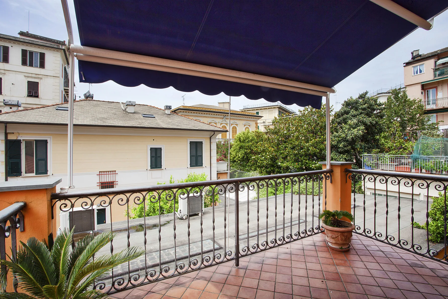 Una terraza de baldosas con toldo oscuro da a edificios cercanos y vegetación.