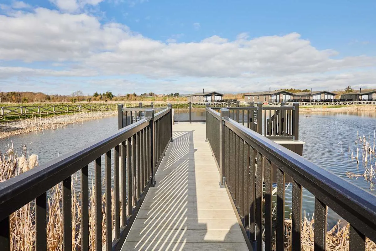 A wooden boardwalk with dark railings crosses a pond toward distant buildings.