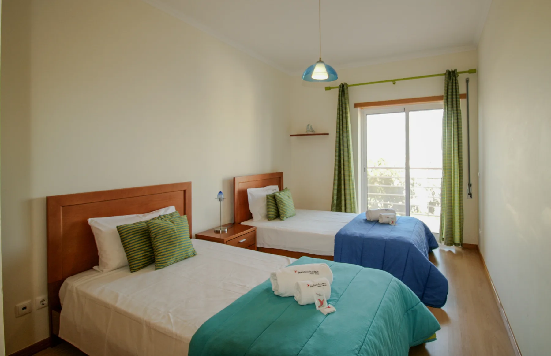 A bedroom with two beds, teal bedding, and a glass door opening onto a balcony.