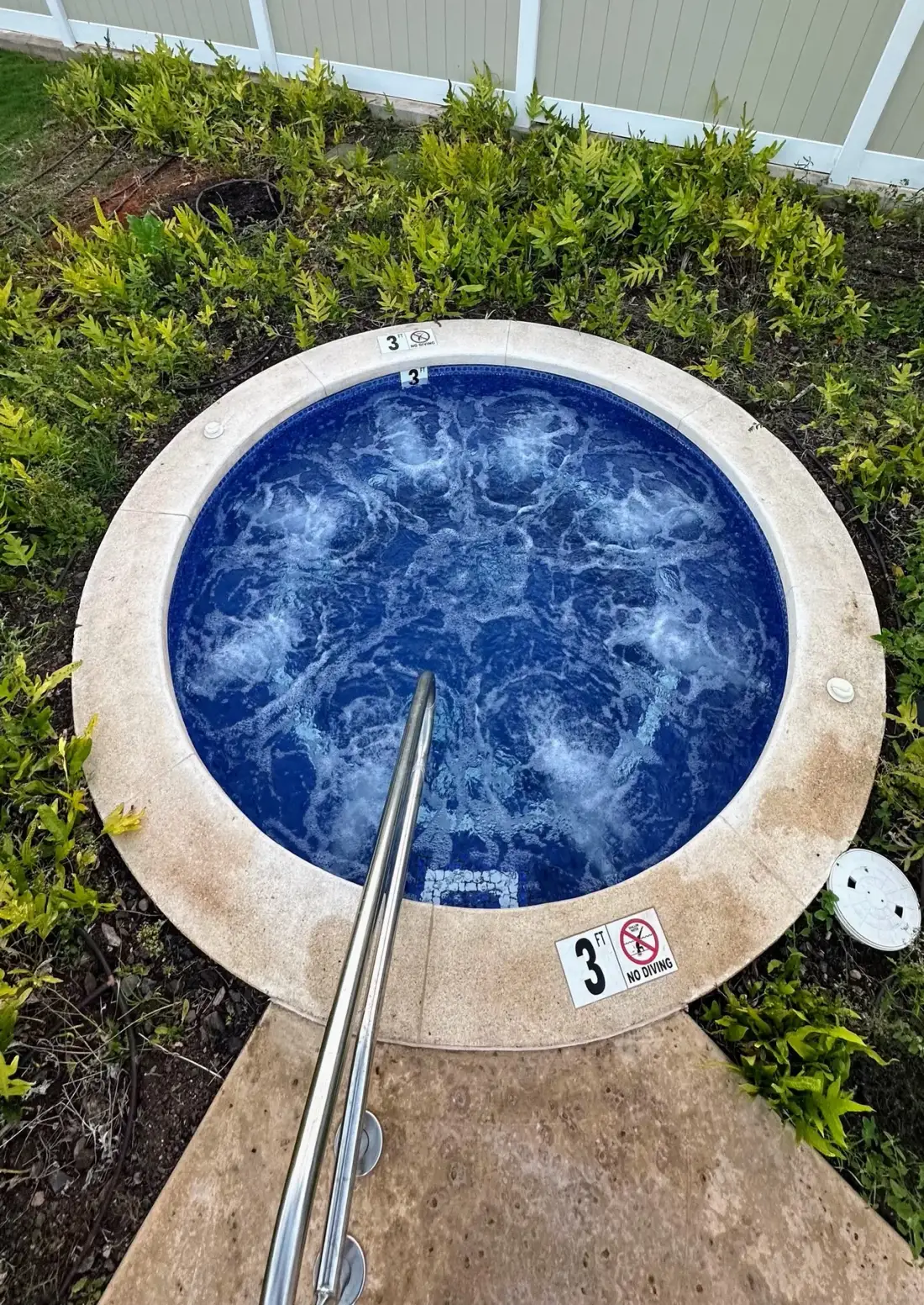 Un jacuzzi circular al aire libre está junto a vegetación baja y una cerca blanca.