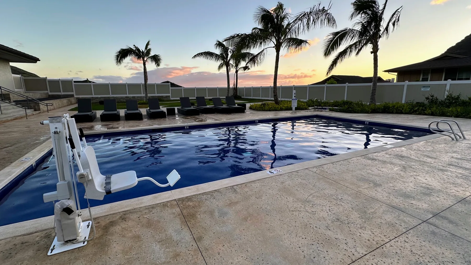 Una piscina al aire libre junto a asientos reclinables, con palmeras y un cielo al atardecer.