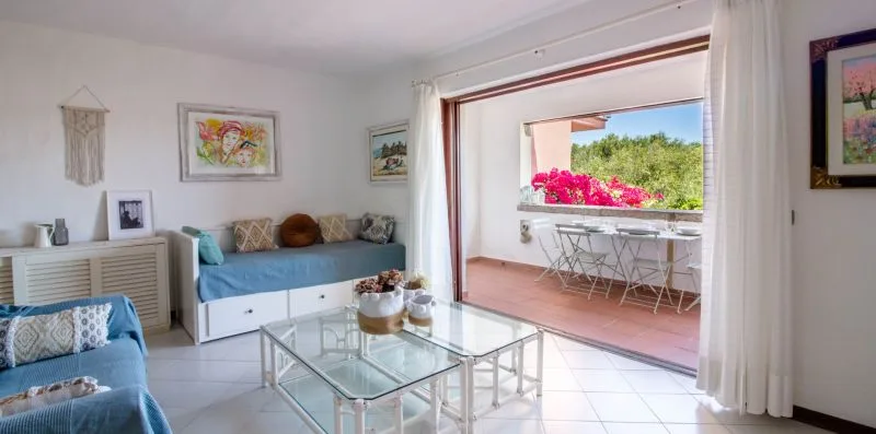 A bright living room opens onto a terrace with outdoor seating and flowering plants.