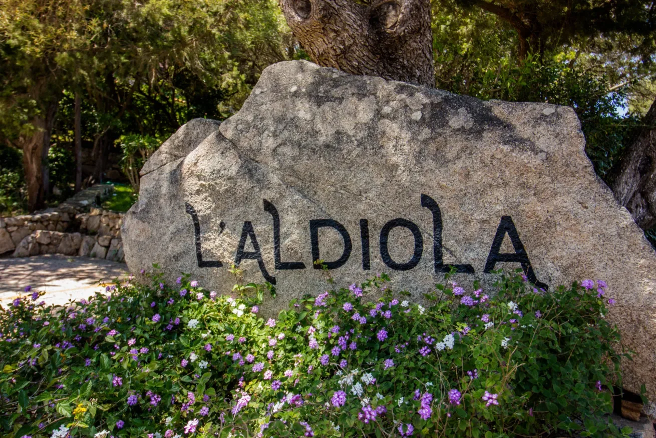 A large stone sign reading “L’ALDIOLA” stands among flowering plants.