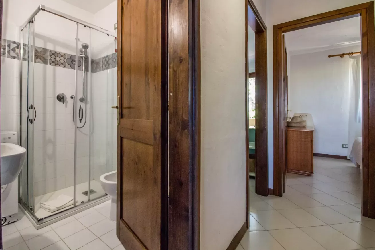 A tiled bathroom with a glass shower and a wooden doorway opening to another room.