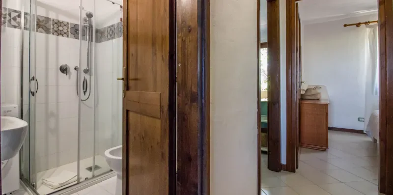 A tiled bathroom with a glass shower and a wooden door opens onto a bedroom.