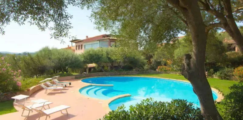 A swimming pool sits beside a sunlit patio with lounge chairs and trees.