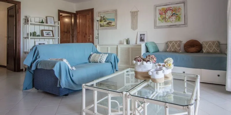 Una sala de estar con asientos cubiertos de azul, mesas de centro de vidrio y cuadros en la pared.
