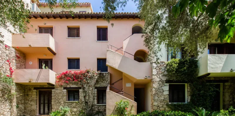 A multi-story residential building with pale walls, balconies, and surrounding greenery.