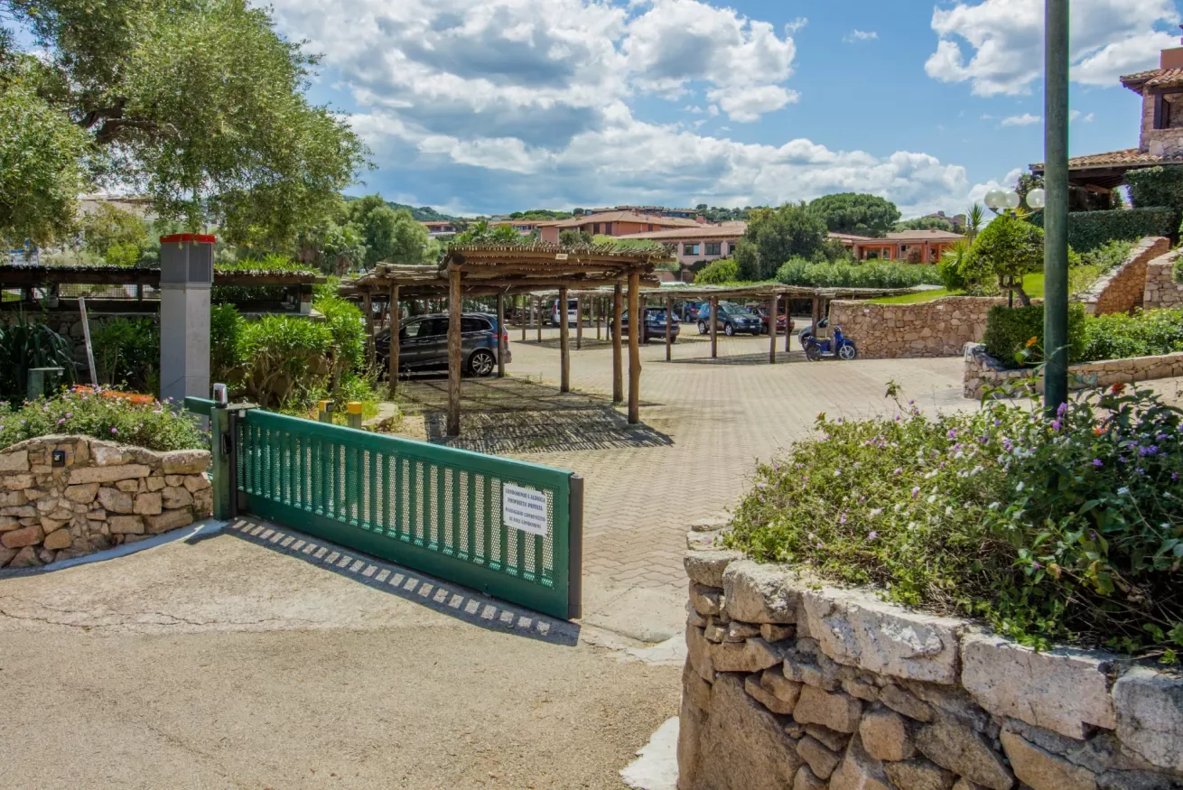 A paved parking area with a wooden carport, greenery, and an open gate.