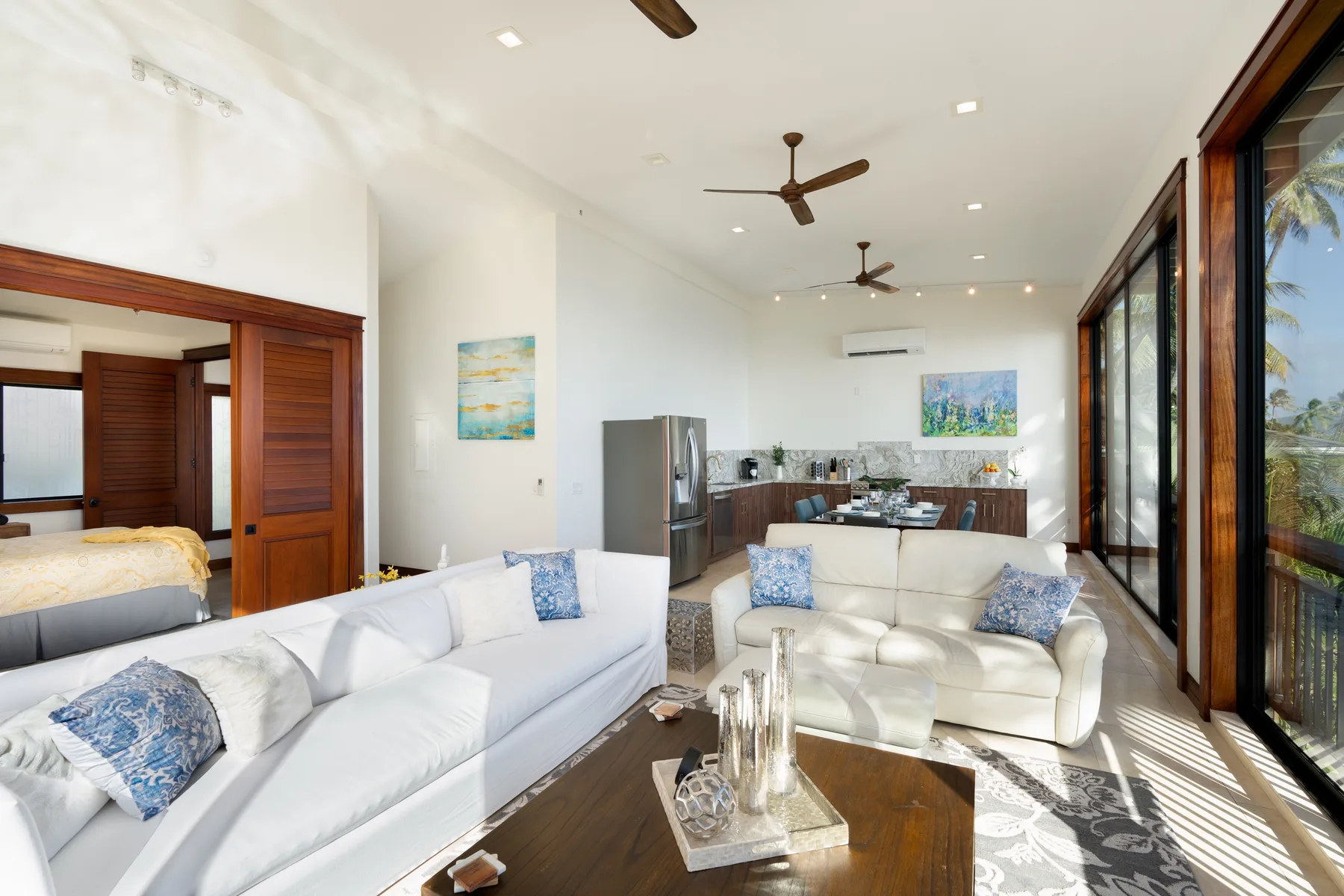 A bright open-plan living room with white seating beside a kitchen and large windows.