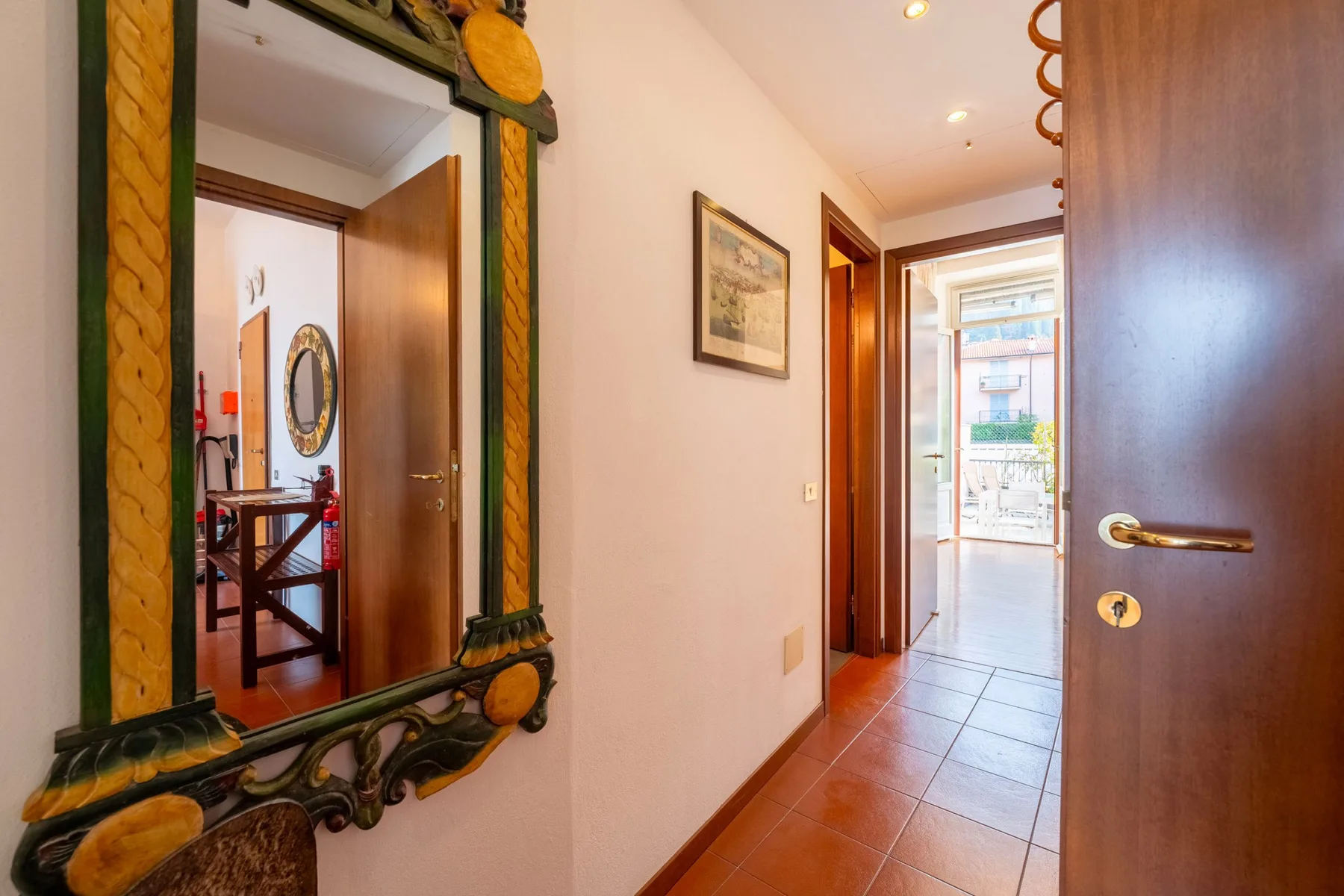Un pasillo con suelo de baldosas, puertas abiertas, un espejo y una ventana luminosa al fondo.