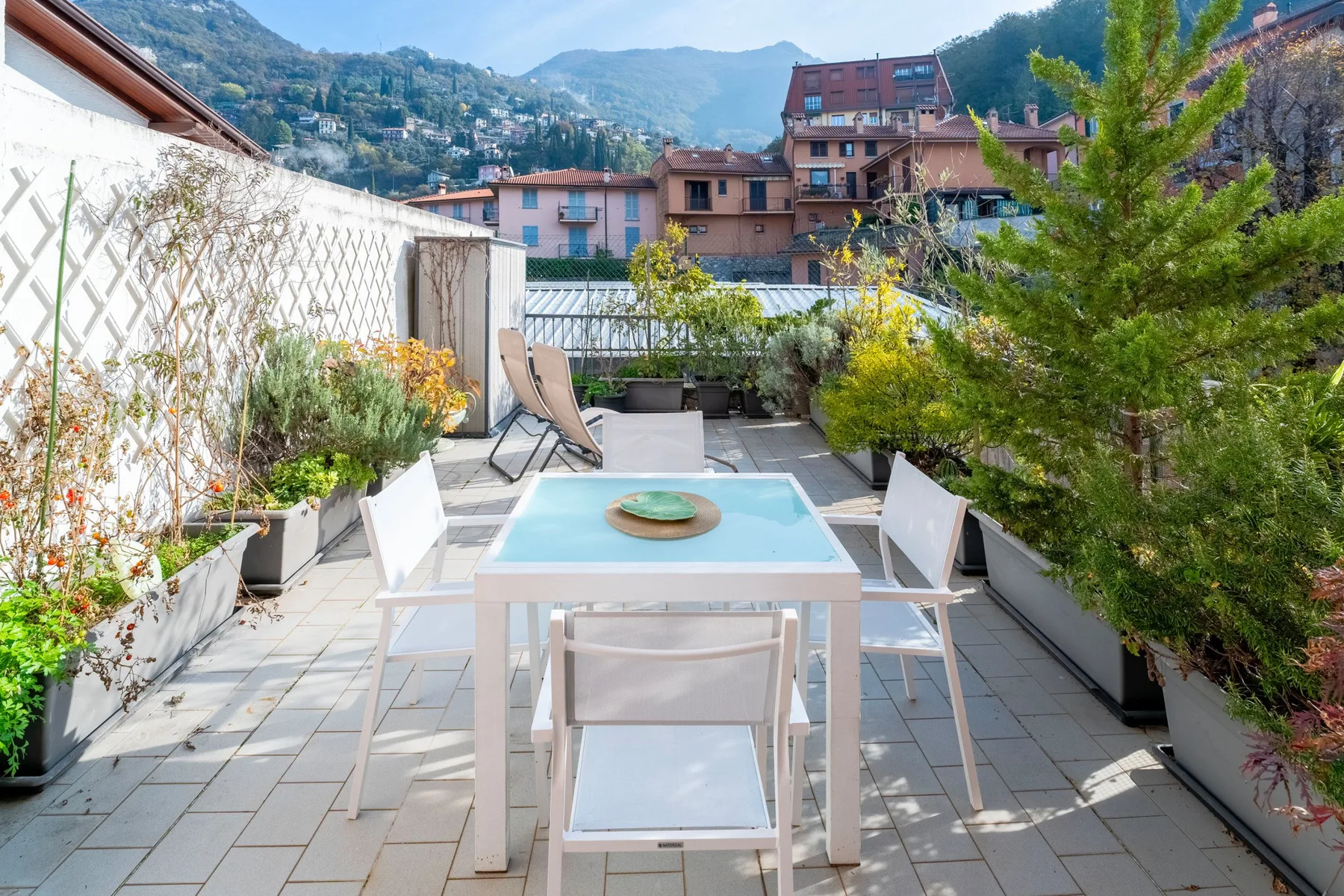 Una terraza exterior embaldosada tiene una mesa de comedor, sillas y plantas en macetas.