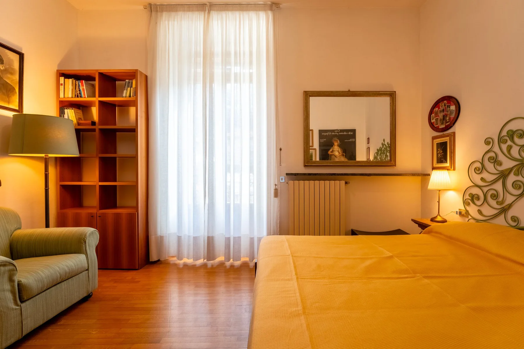 Un dormitorio con una cama, un sillón, una estantería y una ventana alta con cortinas.