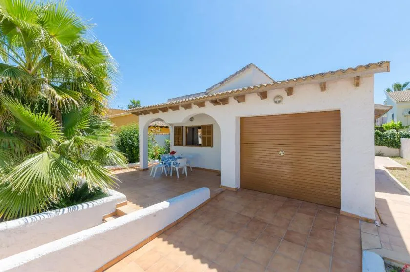 Exterior de una casa blanca con puerta de garaje y asientos en un patio sombreado.