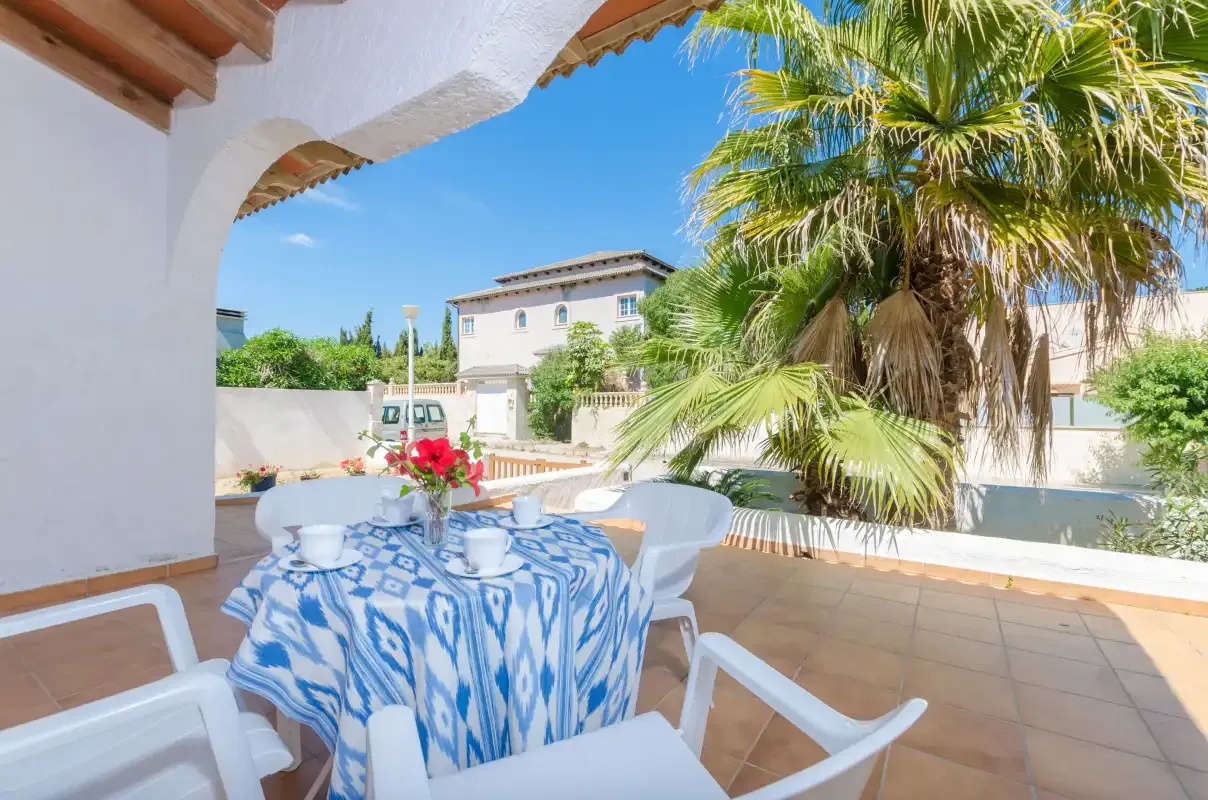 Un patio soleado tiene una mesa con mantel estampado, asientos blancos y una palmera.