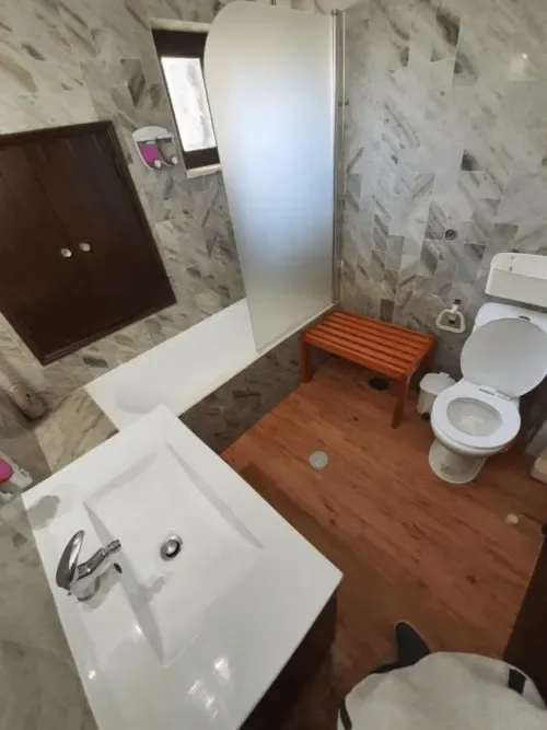 Baño con azulejos de aspecto marmóreo, lavabo blanco, inodoro, bañera y suelo de madera.