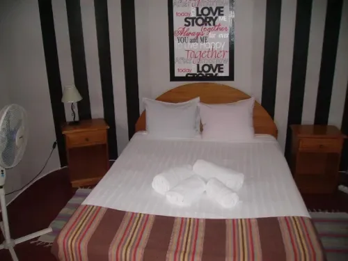 Habitación doble con cama de madera, paredes a rayas, ventilador, mesas de noche y toallas.