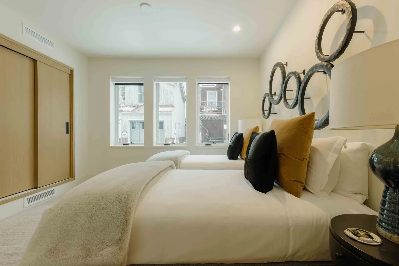 Un dormitorio luminoso con una cama bien tendida y ventanas que dan a edificios cercanos.