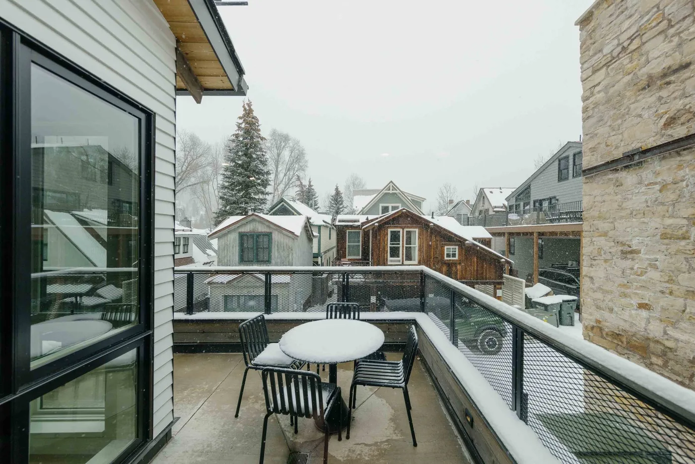 Un patio exterior cubierto de nieve tiene una mesa redonda y asientos junto a una barandilla de vidrio.