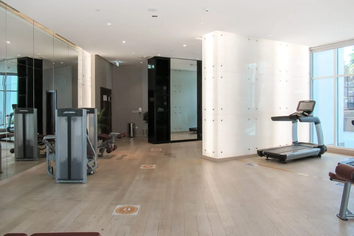 Un gimnasio con máquinas de ejercicio y una cinta junto a grandes ventanas.