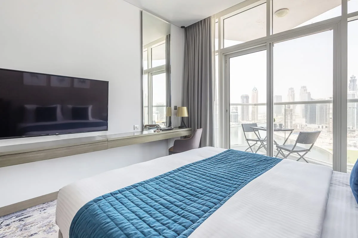 Un dormitorio luminoso con una cama junto a ventanas de piso a techo y un televisor.