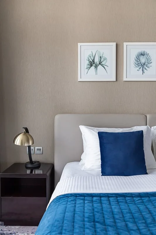 Un dormitorio con una cama bien tendida, un cojín azul y láminas botánicas enmarcadas.