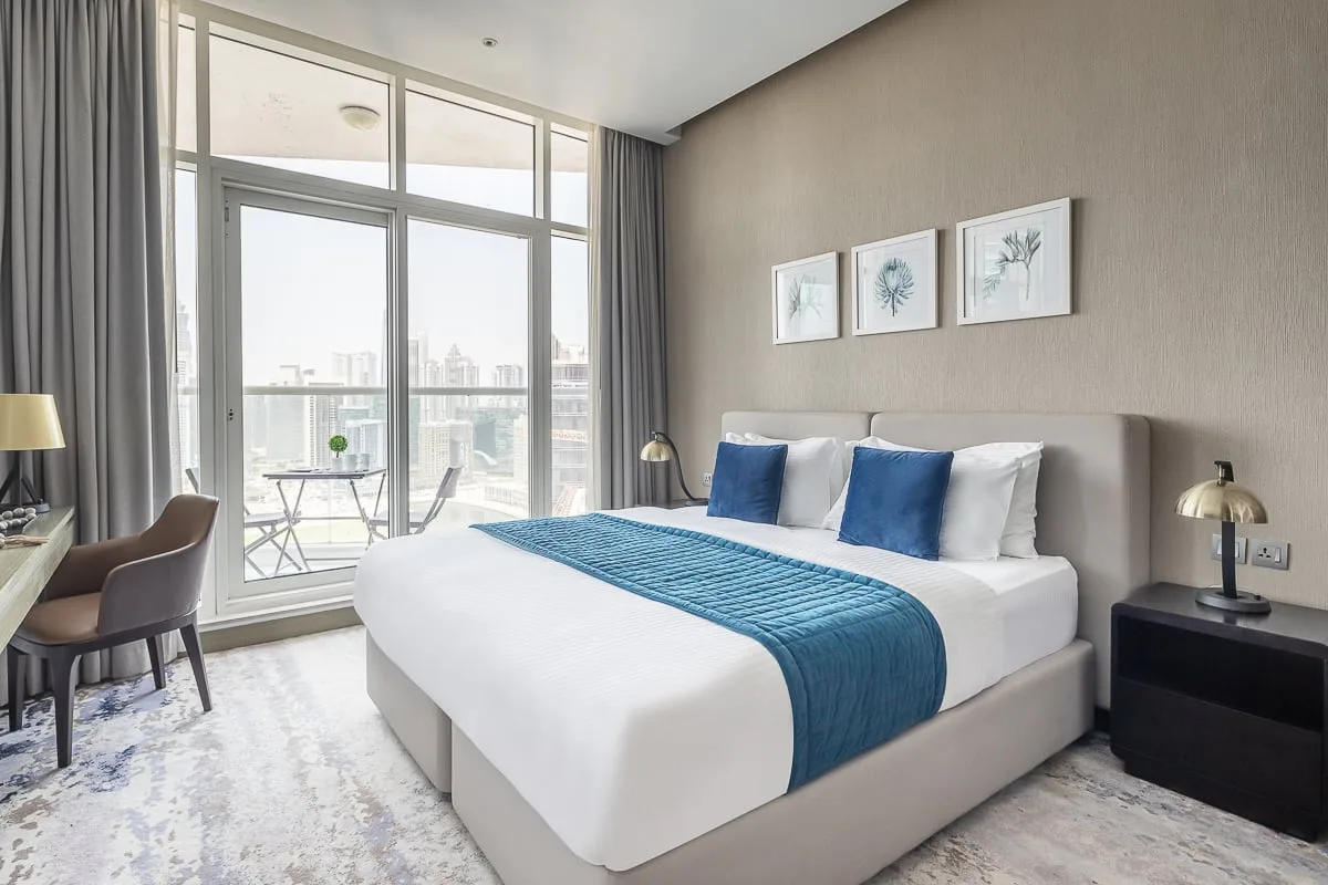 Un dormitorio luminoso con una cama, ropa de cama azul y grandes ventanales hacia un balcón.