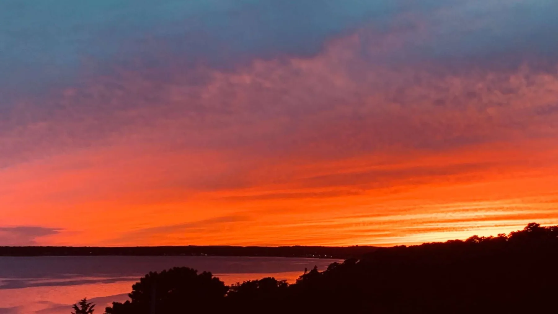 Un atardecer intenso tiñe el cielo sobre un cuerpo de agua tranquilo y una costa oscura.