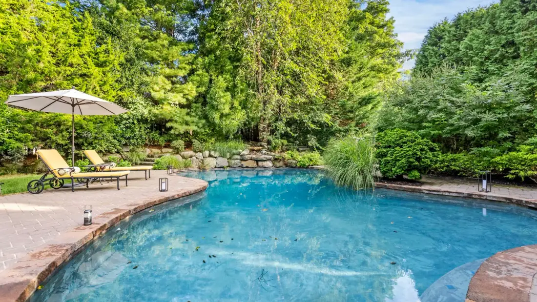 Una piscina al aire libre rodeada de una terraza pavimentada y vegetación frondosa.