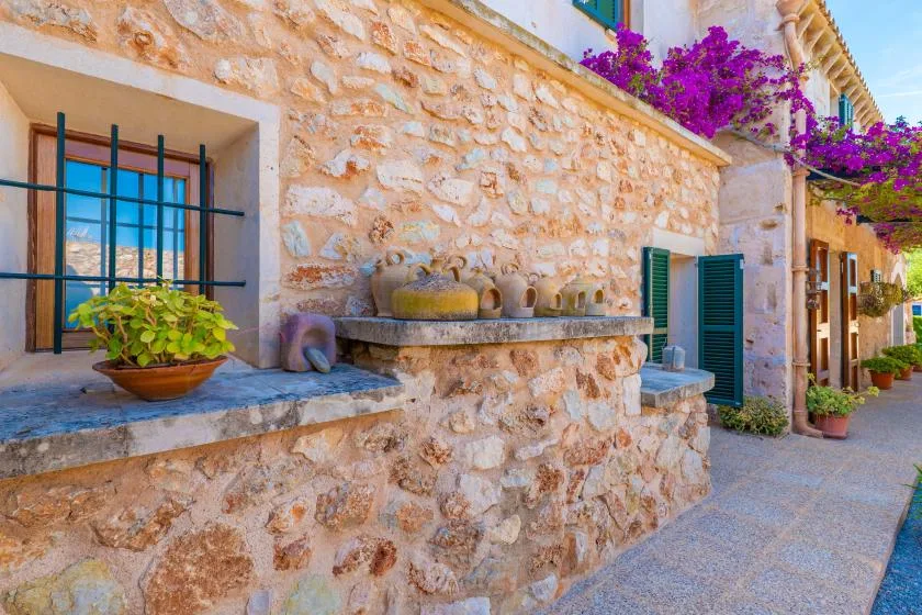 Fachada de piedra con una ventana enrejada, jardineras y un camino pavimentado.
