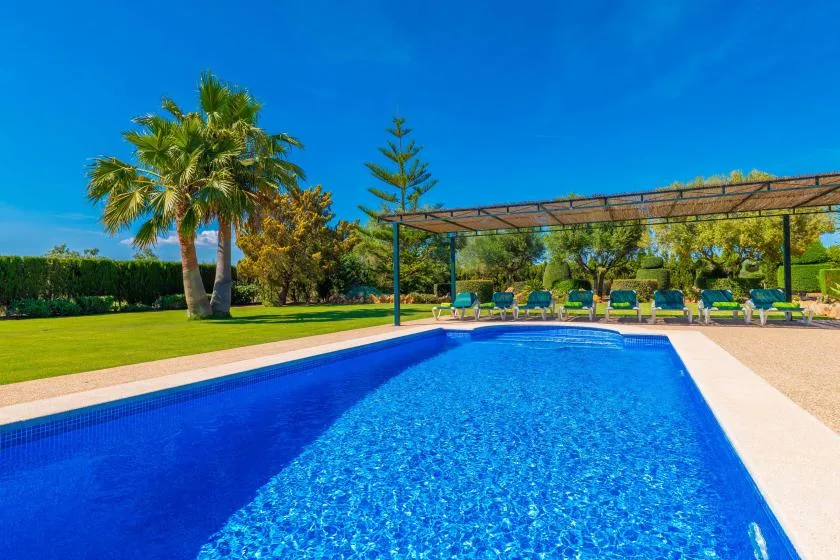 Una piscina al aire libre junto a un césped verde y una zona de asientos a la sombra.