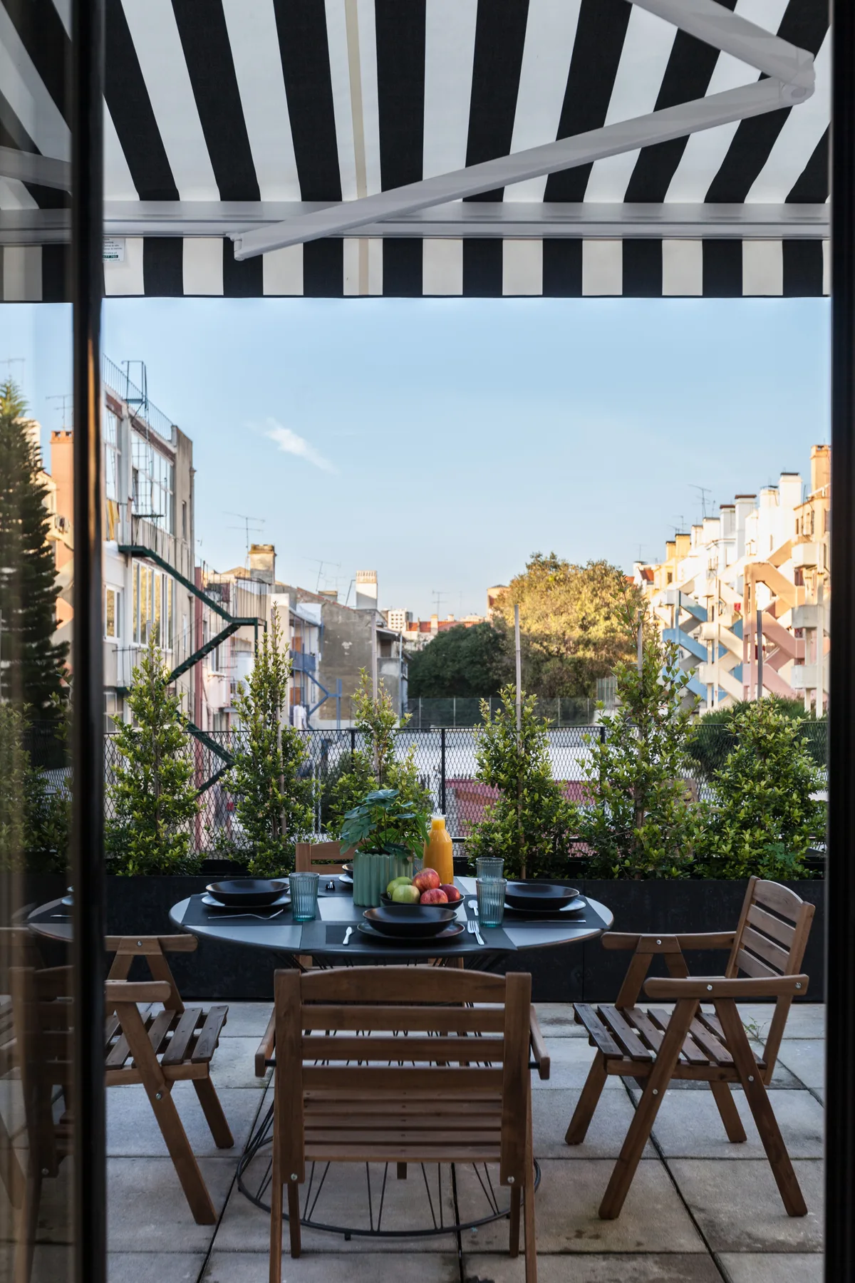 An outdoor dining area with a table and wooden seating overlooks a leafy city street.