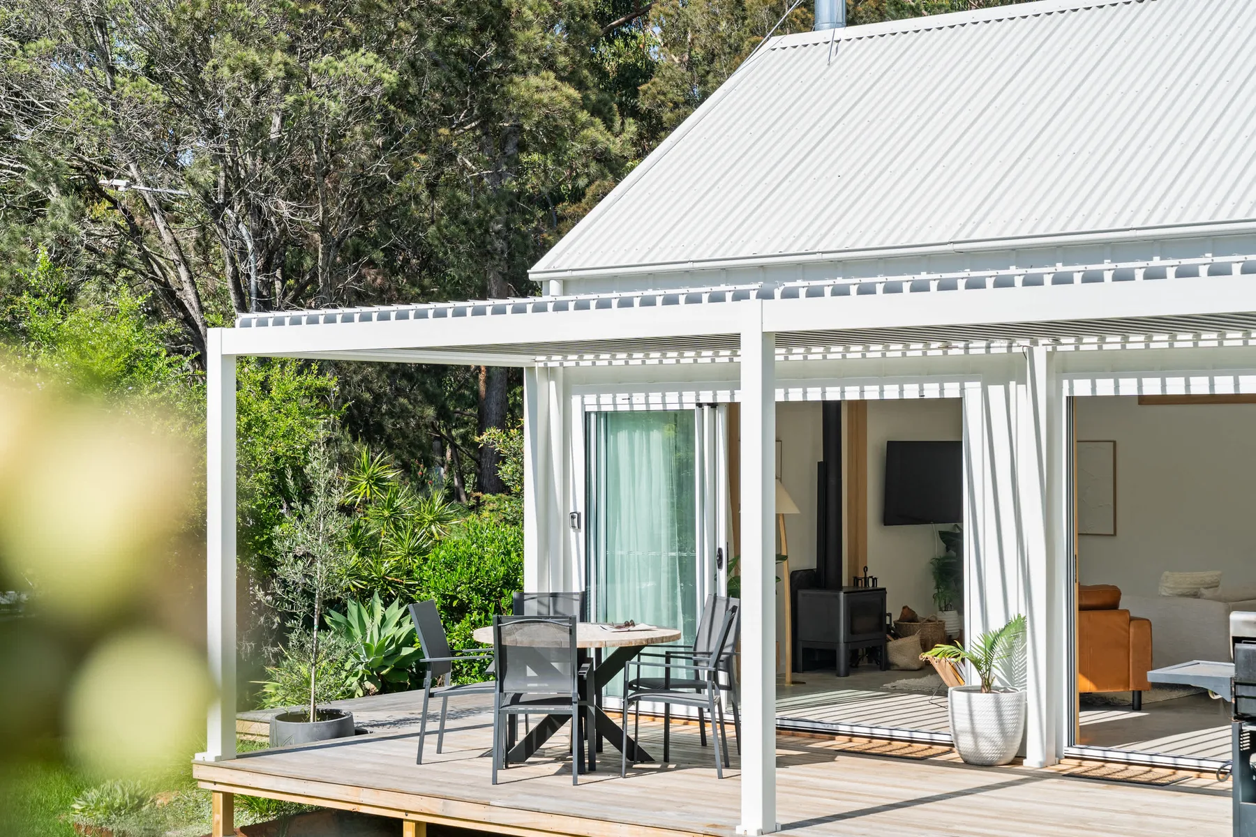 A white house opens onto a wooden deck with outdoor seating and greenery.