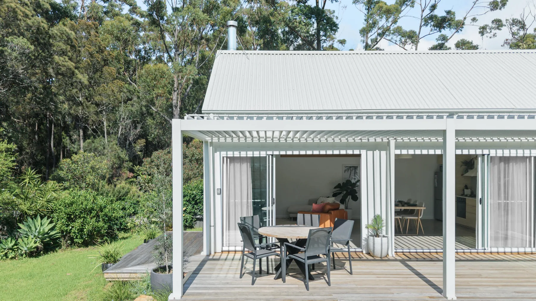 An outdoor patio with seating beside a light-colored house, surrounded by greenery.