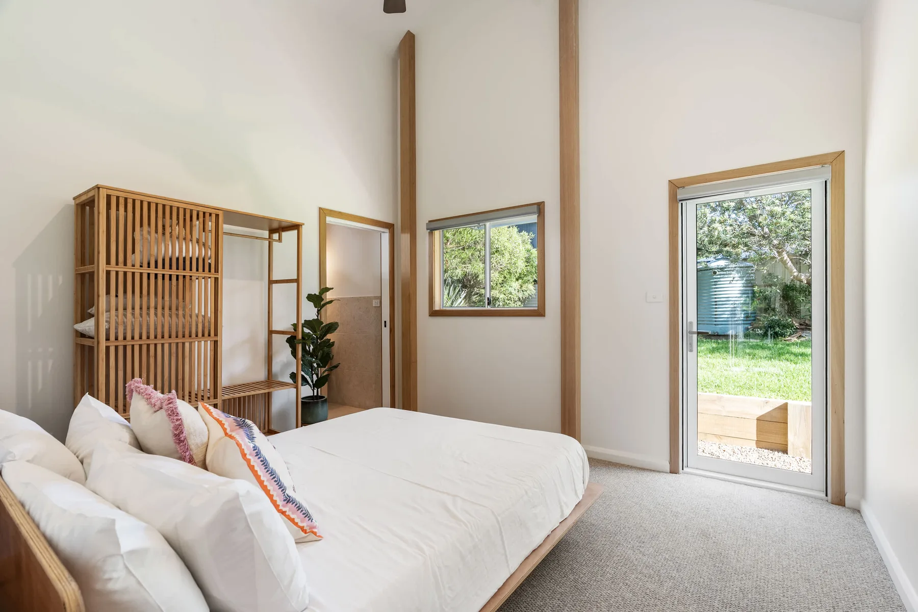 A bright bedroom with a bed, wooden shelving, and a glass door opening to greenery.