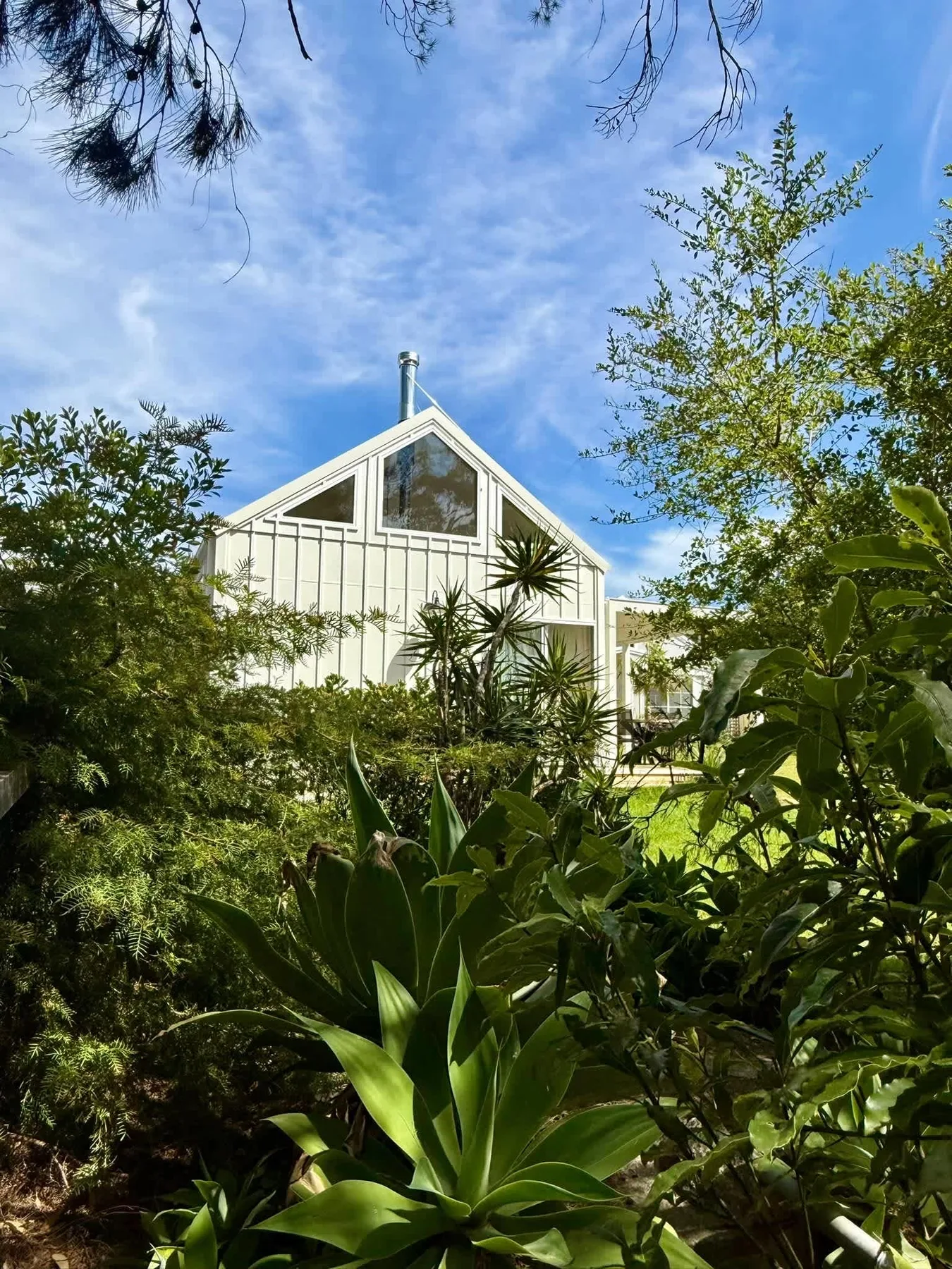 A white gabled building stands among dense greenery beneath a blue sky.