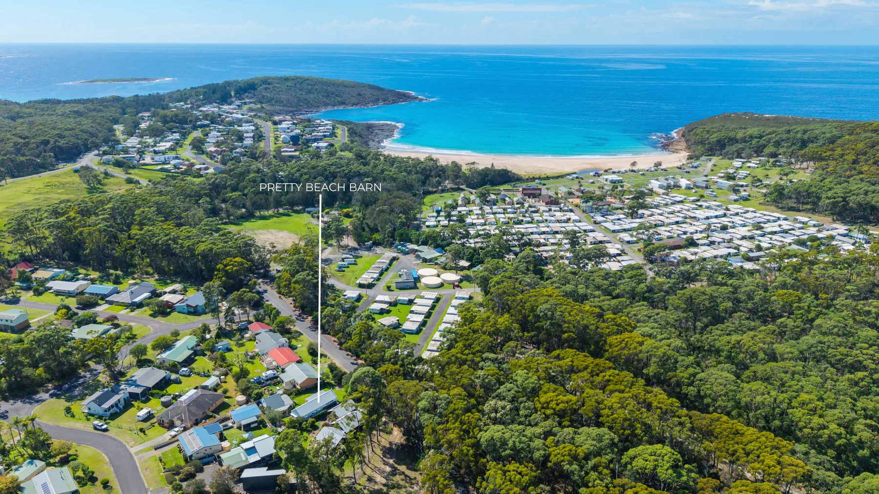 An aerial view shows a coastal settlement nestled between forested hills and a turquoise bay.