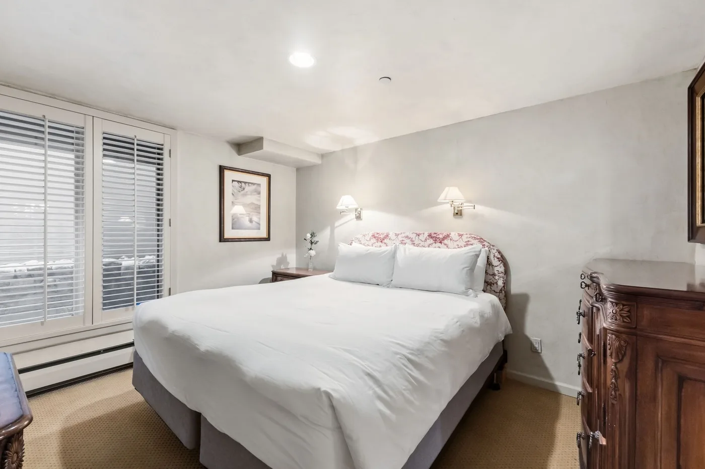 Un dormitorio con una cama bien tendida, lámparas junto a la cama y ventanas con persianas.
