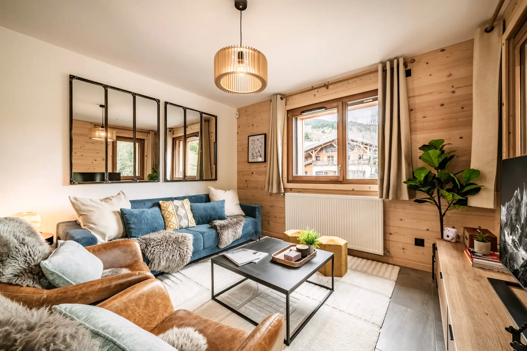 Cozy living room with wooden walls, blue sofa, fur-covered chairs, a small table, and a TV. Large window and balcony door provide natural light.