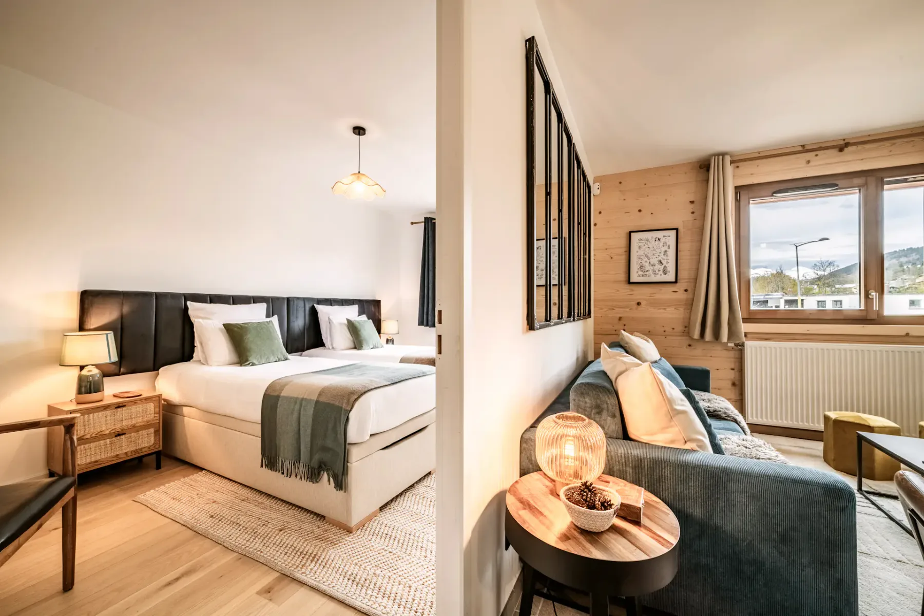 Bright bedroom with two twin beds, green and white bedding, wooden chair, nightstand with lamps, and a window with a view of buildings outside.