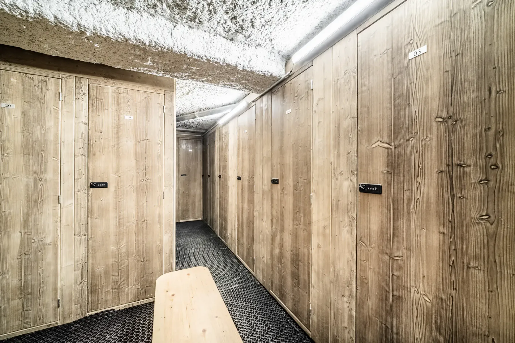 Wooden storage lockers with black number tags line the walls of a room with a textured ceiling and black grid flooring.