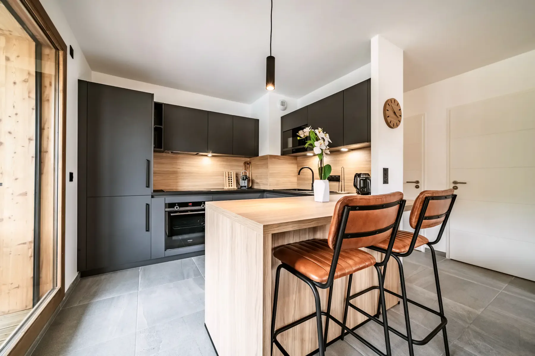 Modern kitchen with dark cabinets, wooden island with two brown stools, pendant light, and wall clock. Bright and spacious with light flooring.