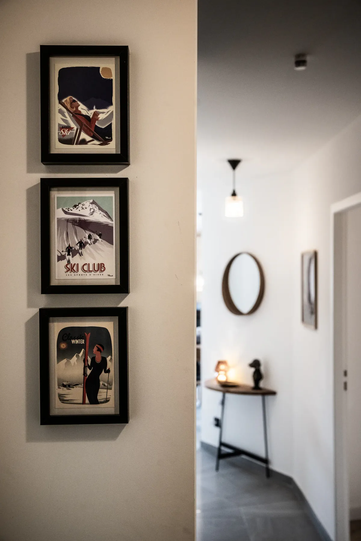 Three framed vintage ski posters on a white wall; hallway with a round mirror and small table in the background.