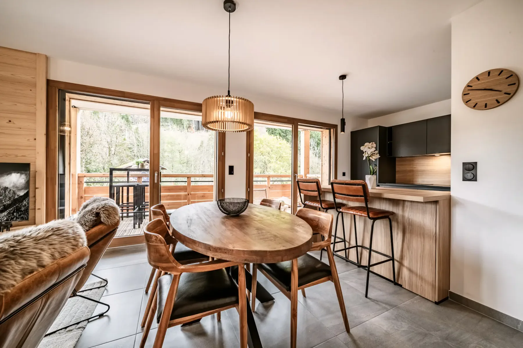 Modern dining area with a round wooden table, leather chairs, and a kitchen island. Large windows overlook a balcony with greenery outside.