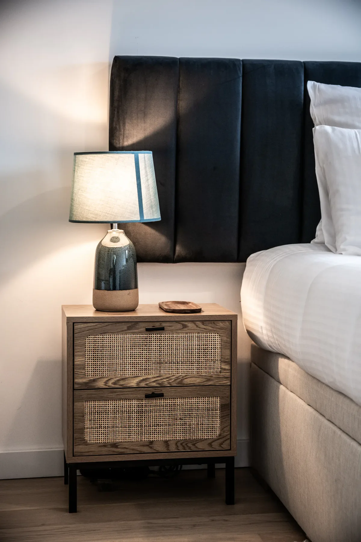 Bright bedroom with two twin beds, green and white bedding, wooden chair, nightstand with lamps, and a window with a view of buildings outside.