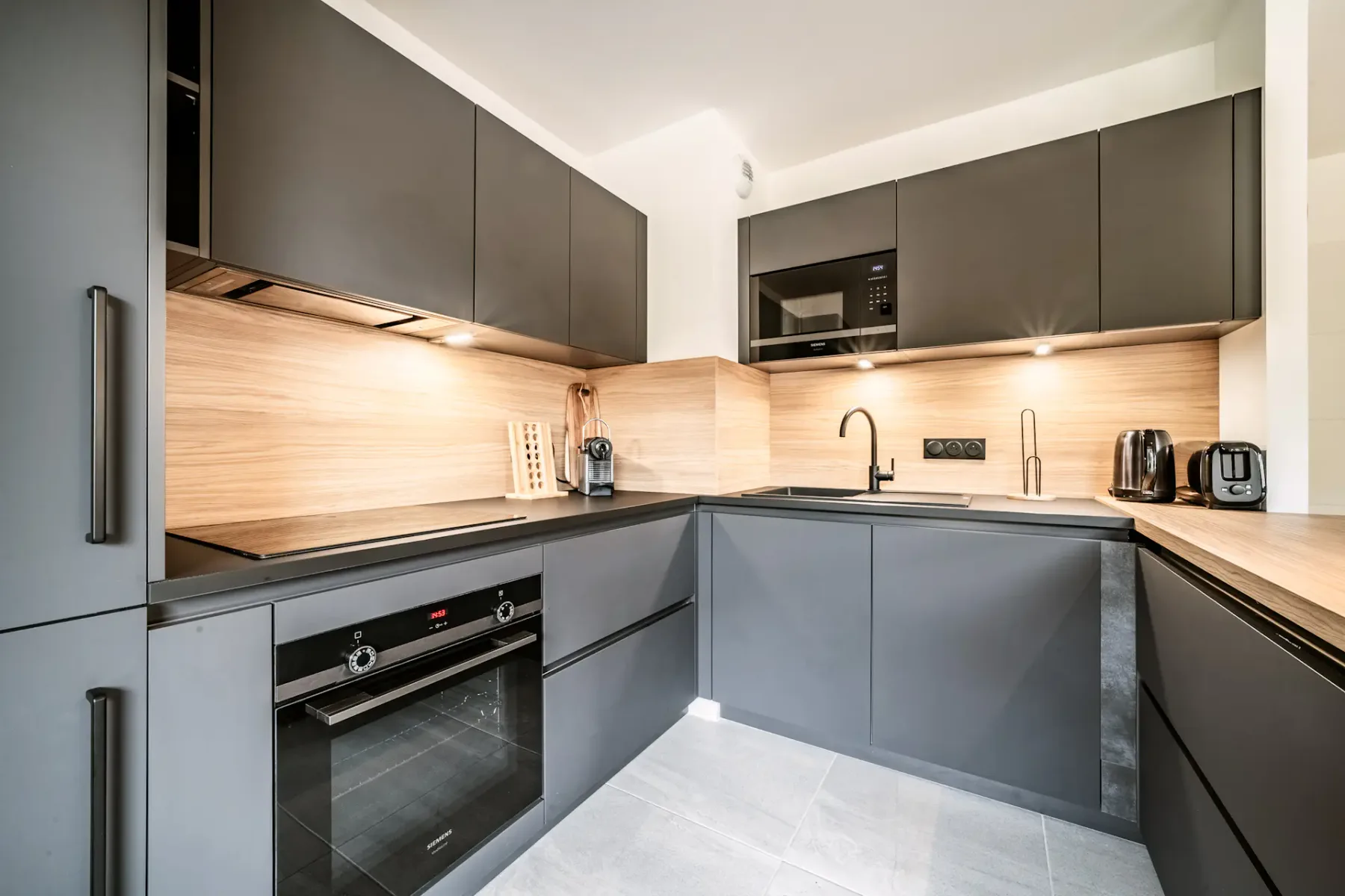 Modern kitchen with dark cabinets, wooden island with two brown stools, pendant light, and wall clock. Bright and spacious with light flooring.