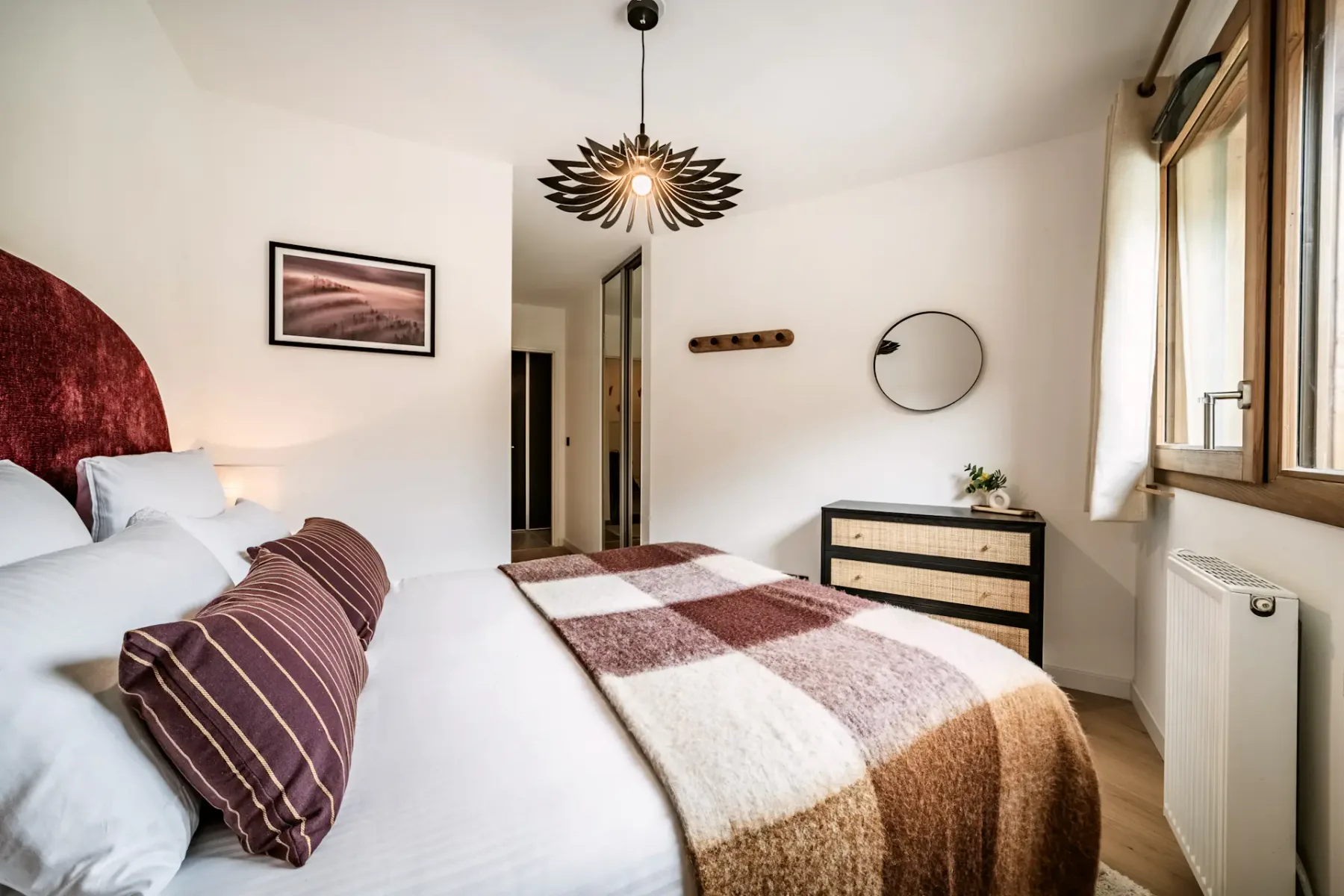 Cozy bedroom with a large bed, plaid blanket, and striped pillows. A unique overhead light fixture and window with curtains brighten the space.