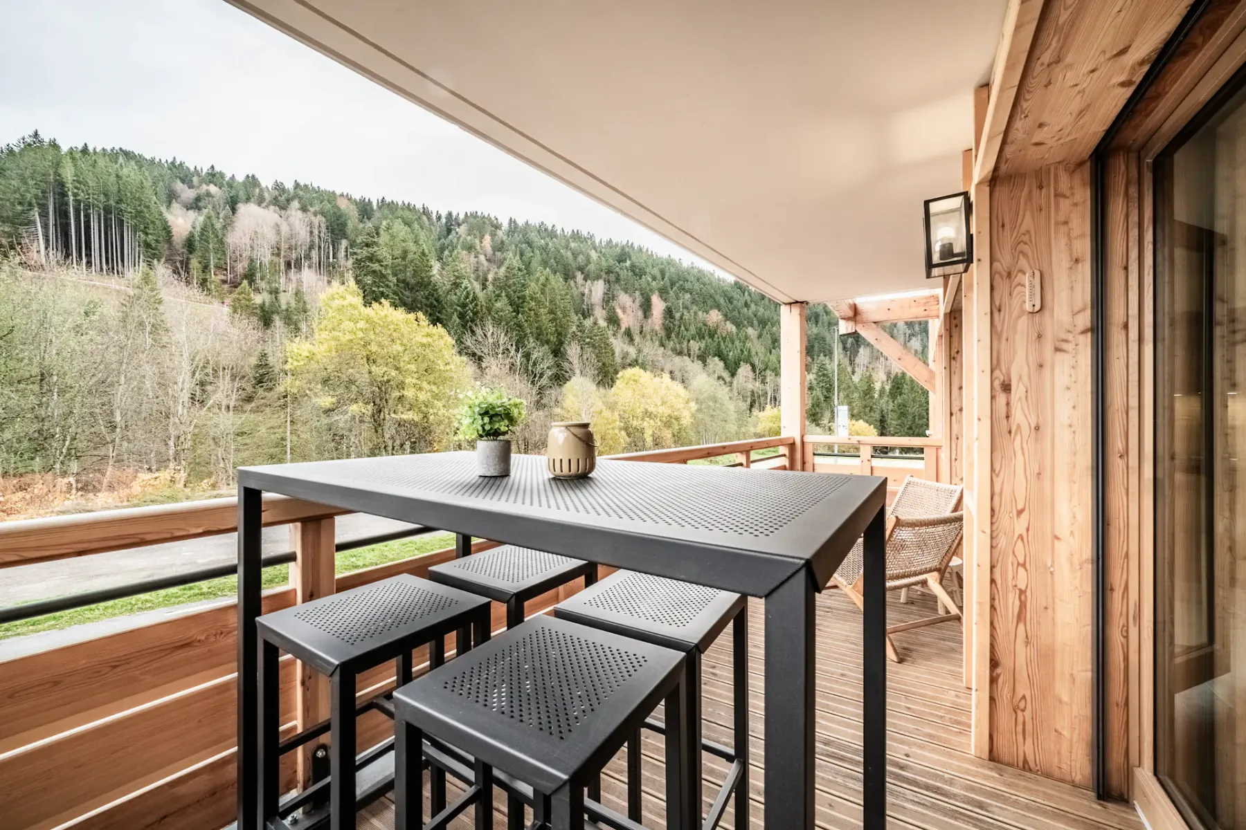 Wooden balcony with a tall black table and stools, overlooking a lush forest landscape under a cloudy sky.